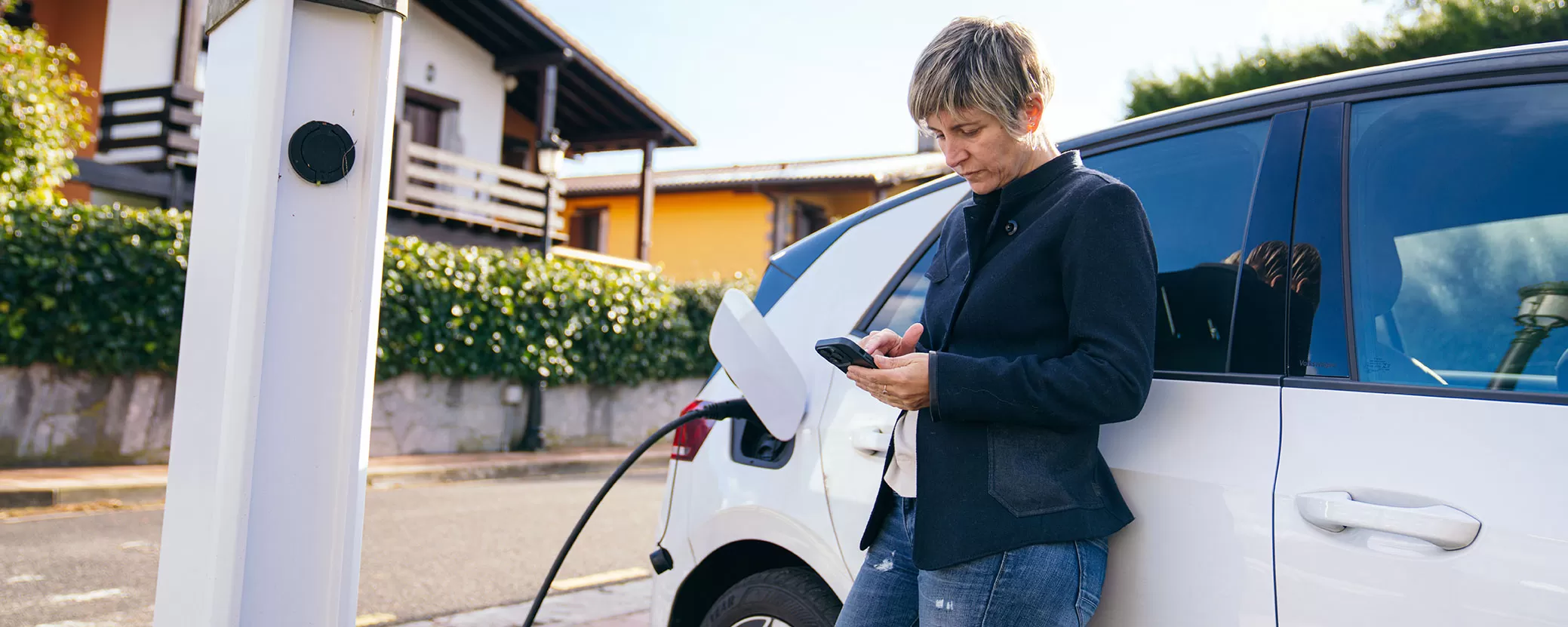 Woman leaning on car checking smartphone as electric vehicle charges